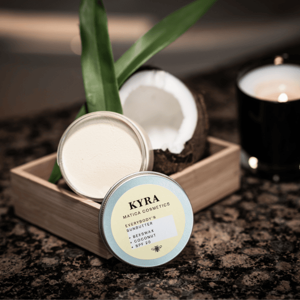 After-Sun Butter Kyra mit Kokosduft, offene Kokosnuss und flackernde Duftkerze auf rustikalem Holz – entspanntes Sommerfeeling trifft natürliche Frische.